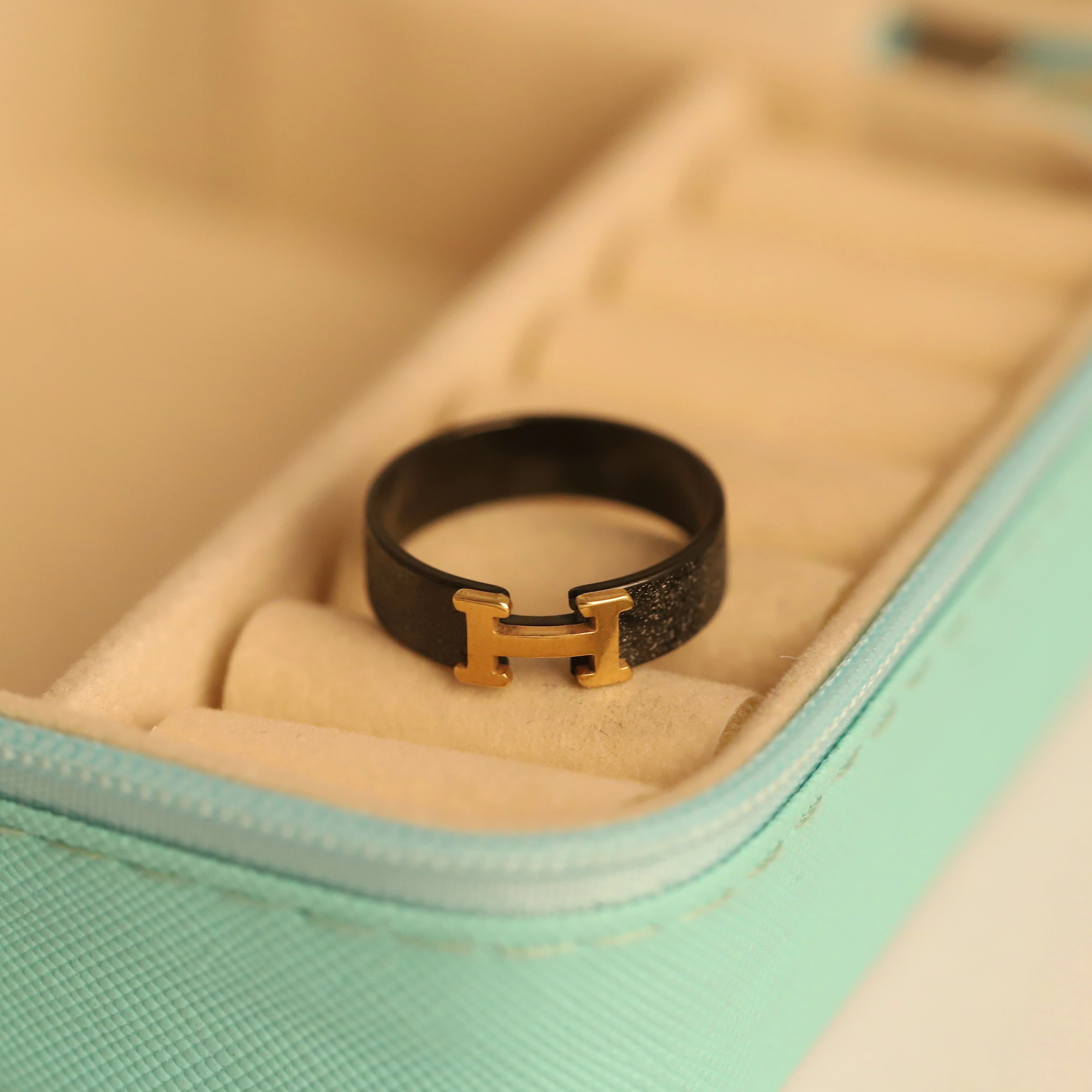 Black ring with gold H logo on a beige cushion in a turquoise jewelry box