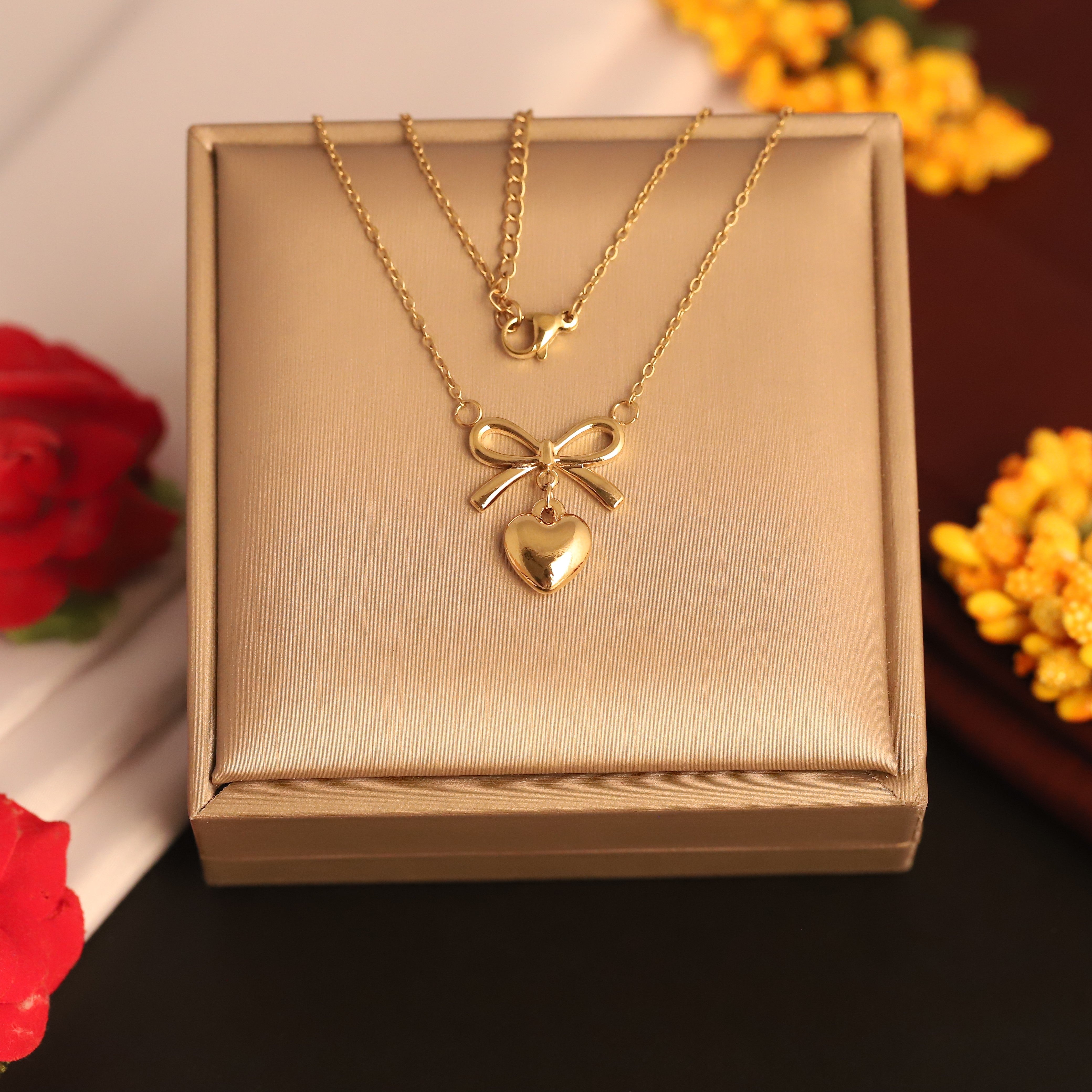 Gold necklace with heart and bow pendant in a box, surrounded by flowers