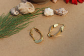 A pair of gold-toned hoop earrings on a brown surface, surrounded by seashells and greenery.